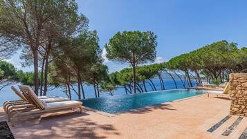An infinity pool with a clear view of the sea, surrounded by trees and lounge chairs.