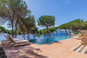 An infinity pool with a clear view of the sea, surrounded by trees and lounge chairs.