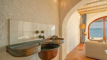 A bathroom with a white tiled wall, a sink with a copper basin, and a view of the sea through an arched doorway.
