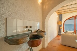 Een badkamer met een witte betegelde muur, een wastafel met een koperen wasbak en uitzicht op zee door een gewelfde deuropening.