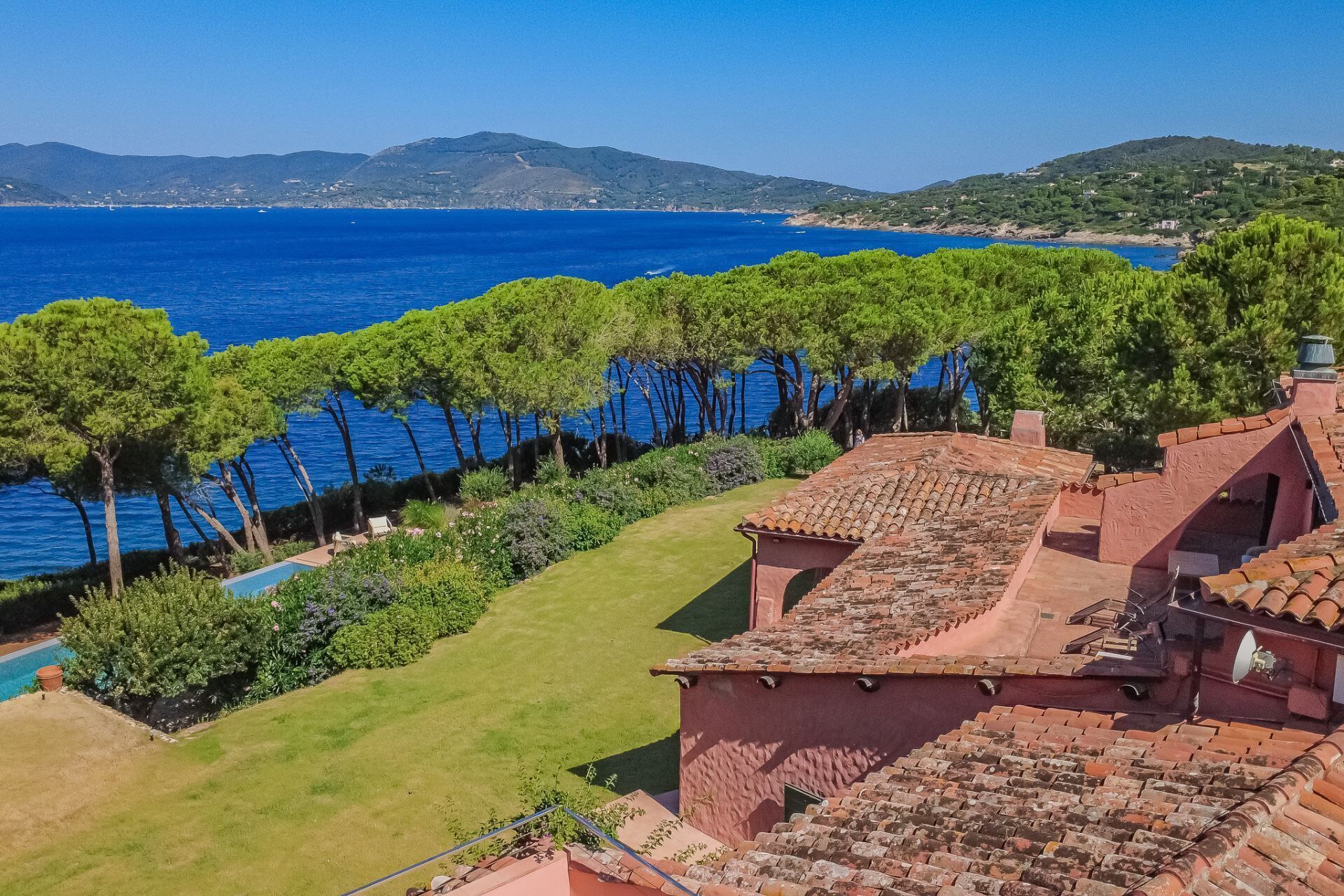 A coastal villa with terracotta roofs, a swimming pool, and a view of the sea and distant hills.