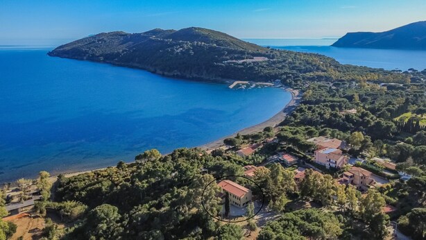 Vista aérea de una zona costera con playa, una pequeña isla y un conjunto de edificios rodeados de vegetación.