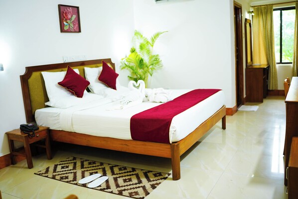 A neatly made bed with a wooden frame, white bedding, and red pillows. A small bedside table with a telephone is visible. A patterned rug is placed under the bed.