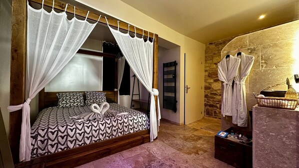 Une chambre à coucher avec un lit, une tête de lit en bois, un mur en pierre et une salle de bain équipée d’un porte-serviettes et d’un panier.