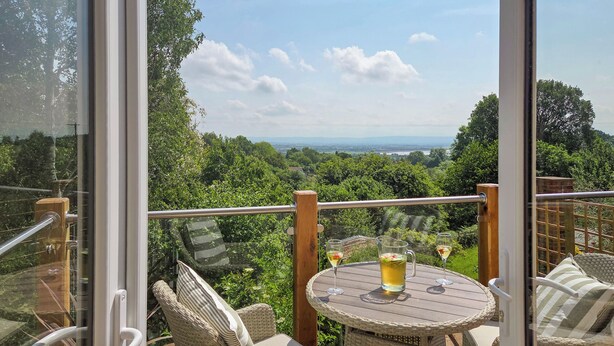 Un balcón con mobiliario de mimbre, una mesa con bebidas y una vista panorámica de vegetación y colinas al lejano.
