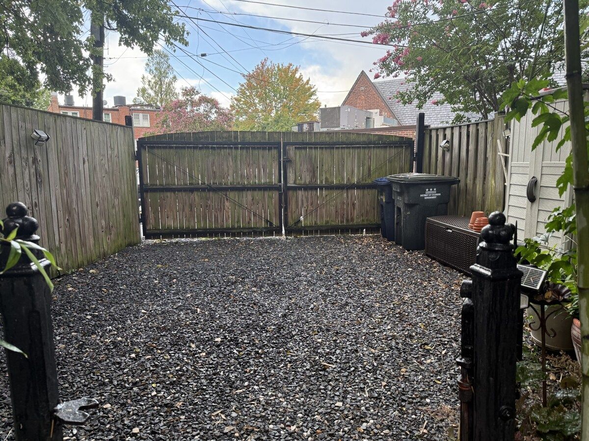Un espace extérieur recouvert de gravier, délimité par une clôture en bois, avec une poubelle noire et un portail en métal.