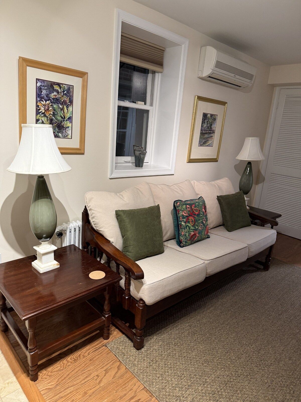 A living room with a sofa, two side tables, a lamp, a radiator, and framed pictures on the wall.