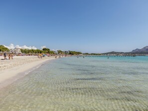 Una playa con aguas poco profundas y cristalinas, un muelle que se adentra en el mar y edificios al fondo.