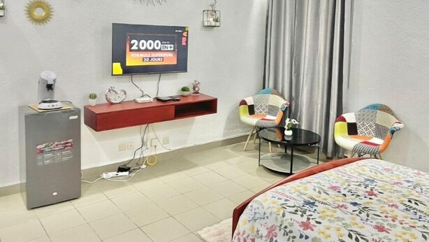 A hotel room with a bed, a TV, a small table, two chairs, a decorative wall clock, and a wall-mounted air conditioner. - Cosy studio at la riviera palmeraie (Abidjan)
