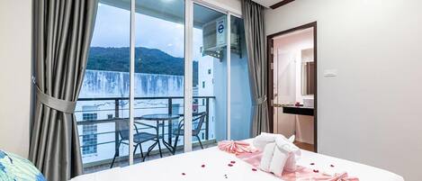 A hotel room with a balcony, a bed with rose petals, and a view of a body of water and hills.