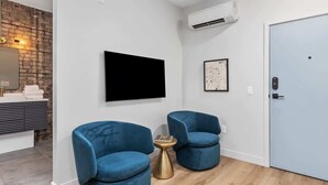 A modern room with a flat-screen TV, two blue armchairs, a small round table, a door, and a brick wall. - 2 Private King Suites - Walk to Nightlife (Grand Rapids)