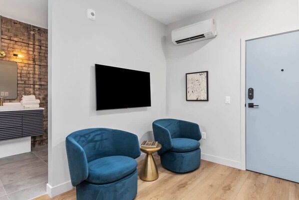 A modern room with a flat-screen TV, two blue armchairs, a small round table, a door, and a brick wall.