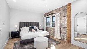 A modern bedroom with a large bed, a round ottoman, a nightstand, and a window with curtains. - 2 Private King Suites - Walk to Nightlife (Grand Rapids)