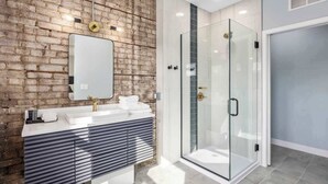A modern bathroom with a brick wall, a glass shower enclosure, and a white vanity with a mirror. - 2 Private King Suites - Downtown GR Getaway (Grand Rapids)