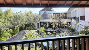 A view from a balcony overlooking a pool area with a thatched-roof structure, surrounded by lush greenery and a building with multiple balconies.