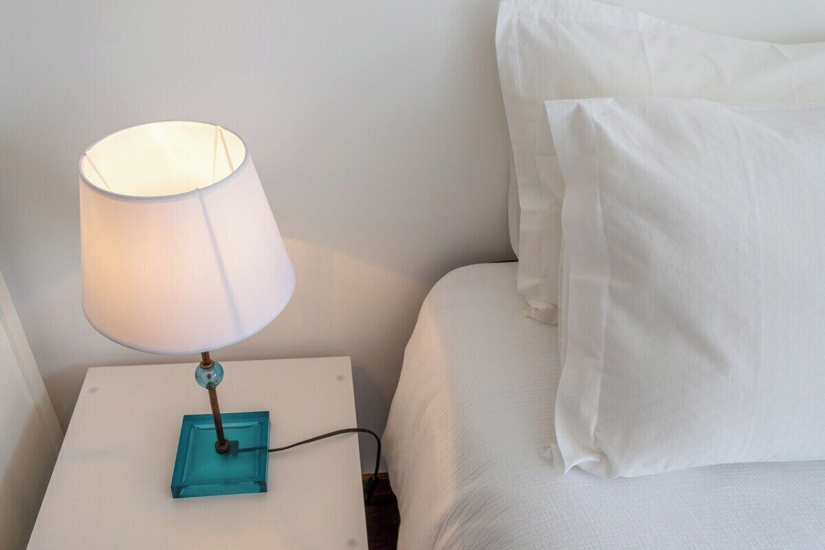 Una cama con ropa blanca y una mesita de noche con una lámpara.