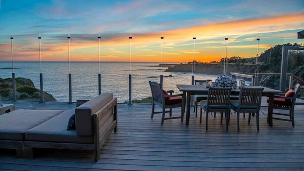 A wooden deck with a table and chairs, overlooking a coastal landscape during sunset. - Dew Point Retreat, by Vinifera Homes (Gualala)