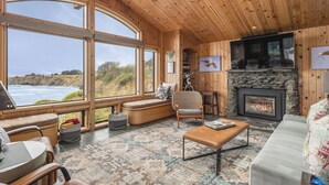 A cozy living room with large windows overlooking the ocean, a stone fireplace, and comfortable seating. - Dew Point Retreat, by Vinifera Homes (Gualala)