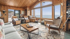 A cozy, wooden-furnished living room with large windows offering ocean views, a comfortable seating area, and a bookshelf. - Dew Point Retreat, by Vinifera Homes (Gualala)