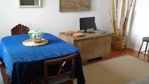 A dining area with a blue tablecloth, wooden chairs, a small table with a decorative item, a television on a wooden stand, and a wall-mounted shelf with a woven basket. - GuestReady - Cheerful Ponta Delgada townhouse (Ponta Delgada)