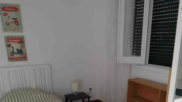 A small room with a single bed, a nightstand, a lamp, and a window with blinds.
