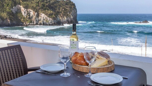 A table set with a bottle of wine, glasses, and a plate of cheese and bread, overlooking a coastal view.