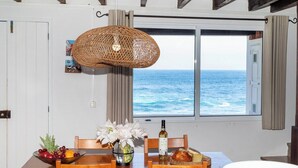 A dining area with a table set for two, a wicker pendant light, a view of the sea through a window, and a bowl of fruit on the table.