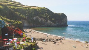 A beach with rocky terrain, clear blue water, and a hilly backdrop.