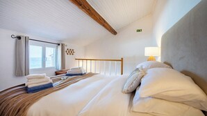 A hotel room with a large bed, a window with curtains, a wooden headboard, a bedside lamp, and a wall-mounted light switch.
