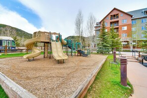A playground with a slide and climbing structures, surrounded by a gravel area and a building with multiple balconies.