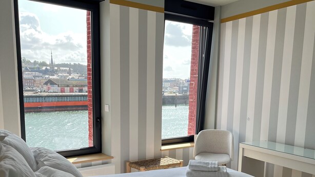 A hotel room with a large bed, a chair, a small table, and a view of a cityscape through a window. - The Touline Apple **** (Fécamp)