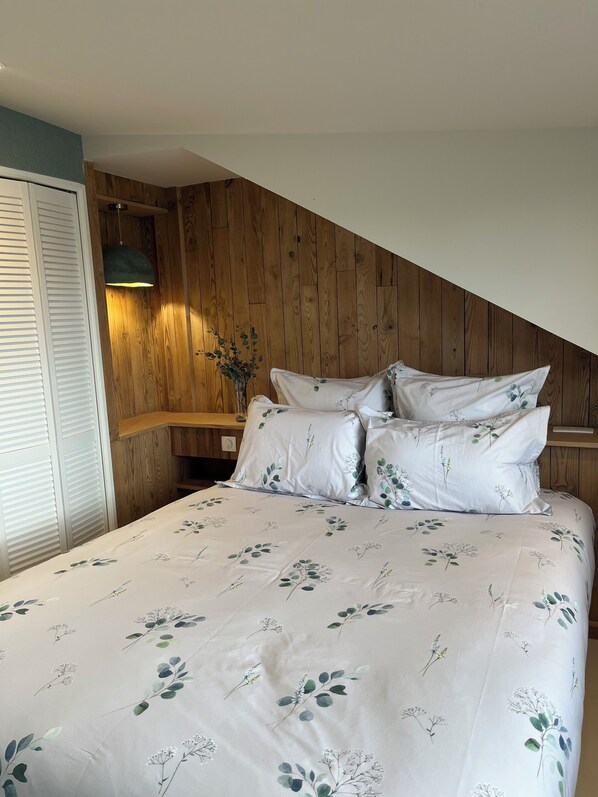 A bedroom with a wooden sloped ceiling, a bed with floral bedding, and a nightstand with a lamp.