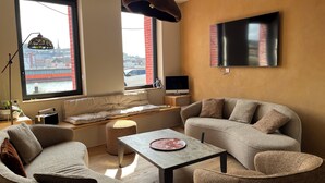 A modern living room with a sofa, armchair, and ottoman, a flat-screen TV, a small table, and a window with a view of a bridge and buildings. - The Touline Apple **** (Fécamp)