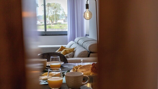 Une table dressée pour le petit-déjeuner avec des assiettes, des verres et une tasse, un canapé avec des coussins et une fenêtre donnant sur un paysage verdoyant.