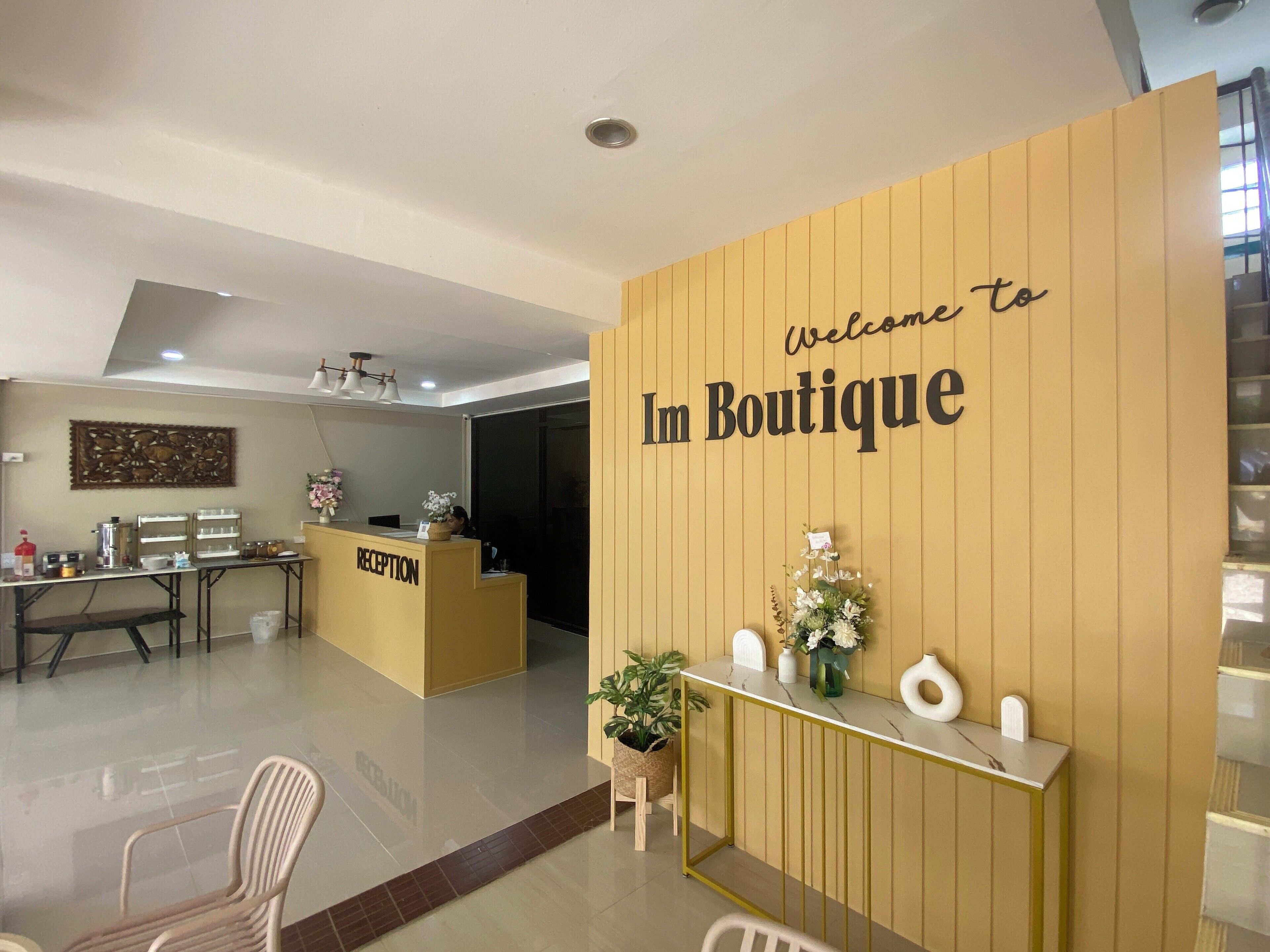 Un área de recepción con un cartel que dice 'Welcome to Im Boutique', un mostrador de recepción, un espacio de descanso con sillas y una pared decorativa con un arreglo floral.