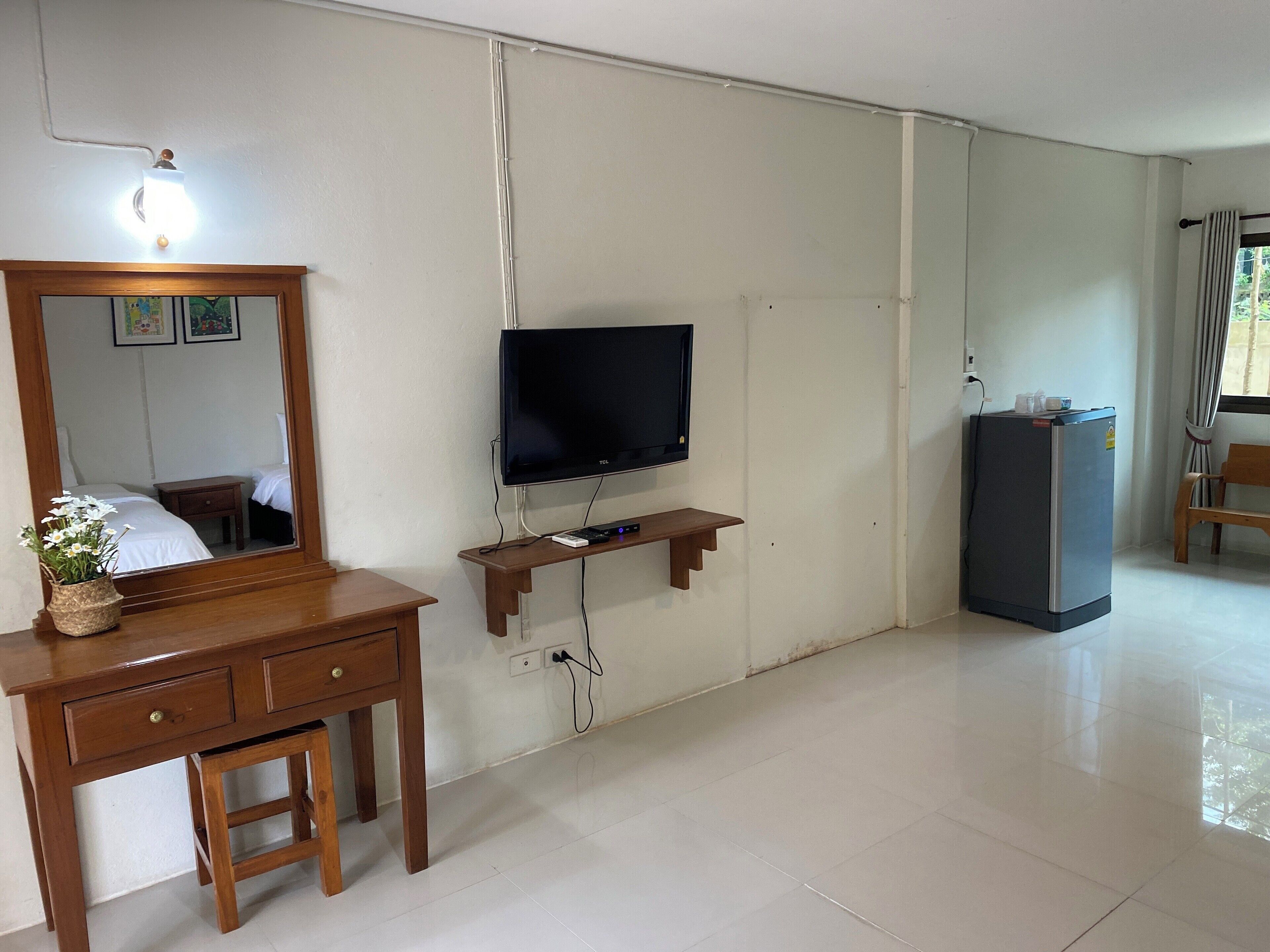 Una habitación con escritorio de madera, espejo, televisor de pantalla plana, refrigerador pequeño y ventana con cortinas.