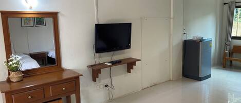Una habitación con escritorio de madera, espejo, televisor de pantalla plana, refrigerador pequeño y ventana con cortinas.