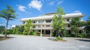 Un hôtel à plusieurs étages, doté d’une entrée centrale et entouré de verdure, avec un ciel dégagé.