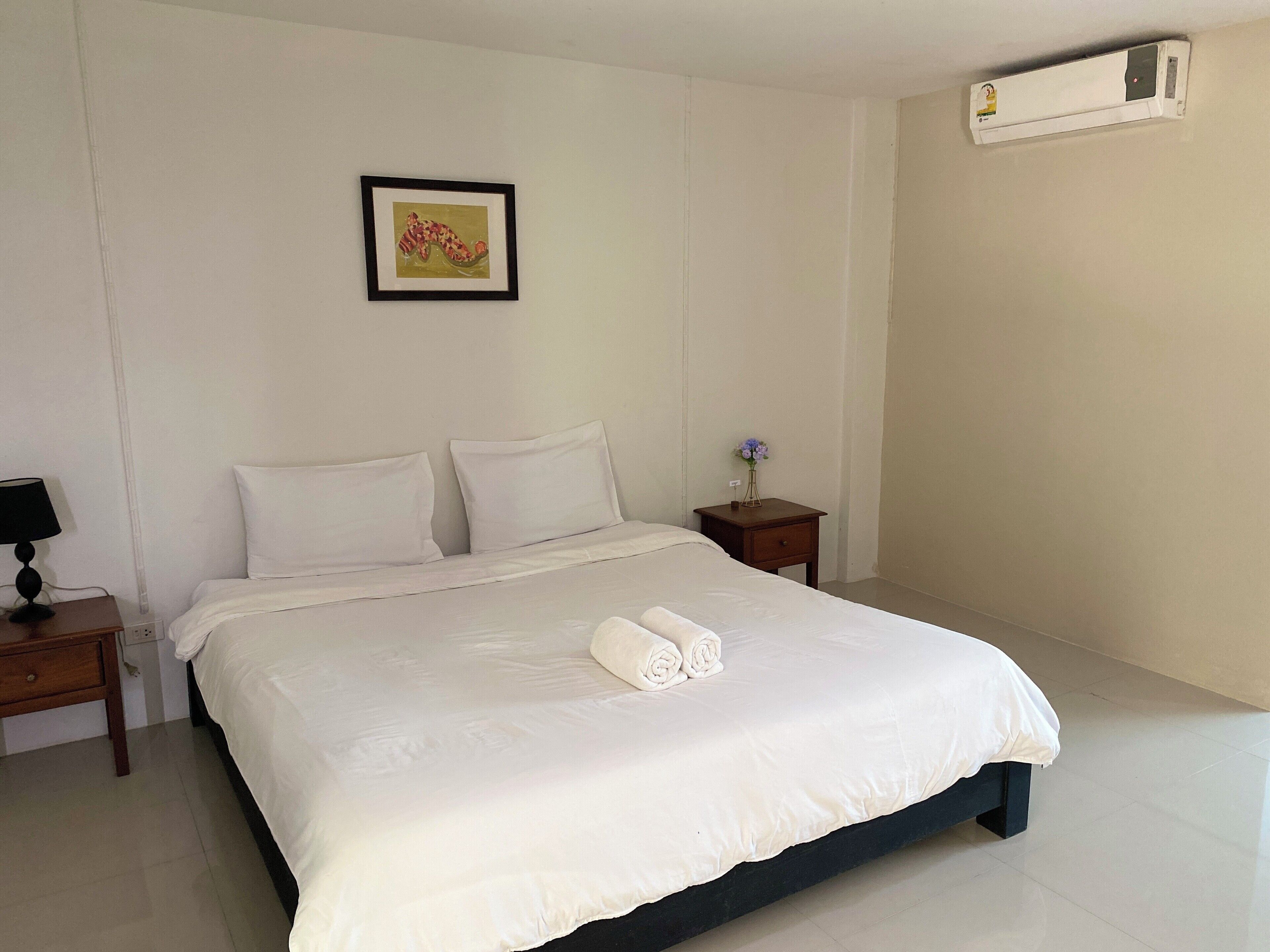 Una cama bien hecha con sábanas blancas y una toalla enrollada, una mesita de noche con una lámpara, un cuadro en la pared y un aire acondicionado instalado en la pared.