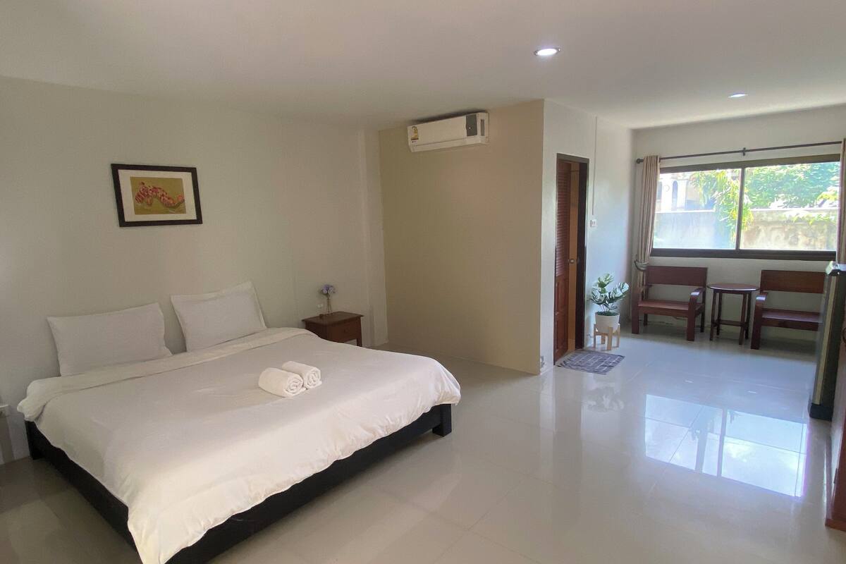 A hotel room with a large bed, a small bedside table with a lamp, a framed picture on the wall, and a window with a view of greenery outside.
