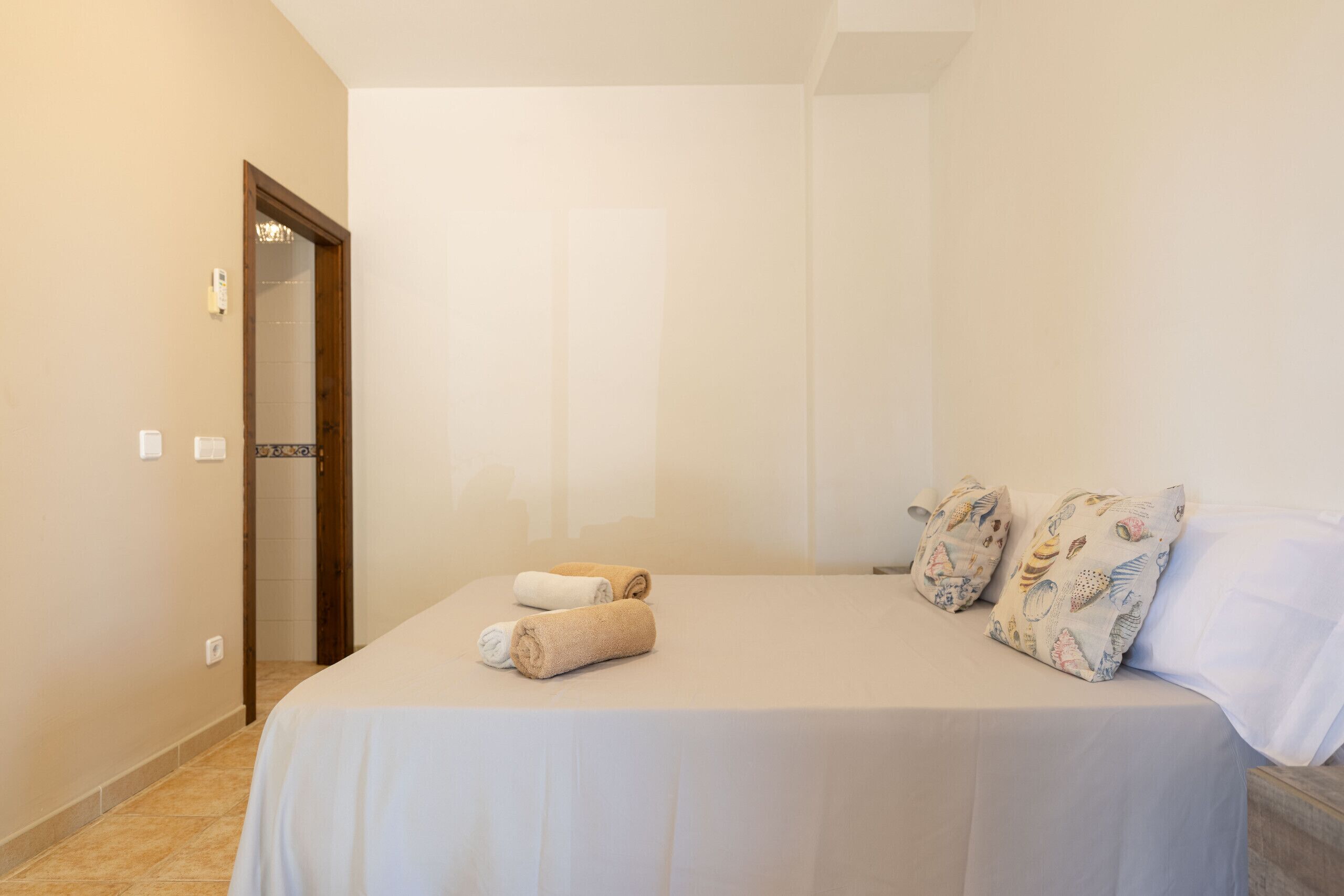 A hotel room with a bed, two pillows, a rolled-up towel, and a door leading to another room.