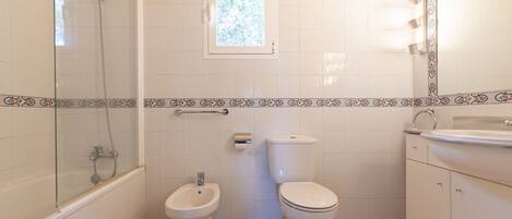 A bathroom with a shower, two toilets, a bidet, a window, and a sink.