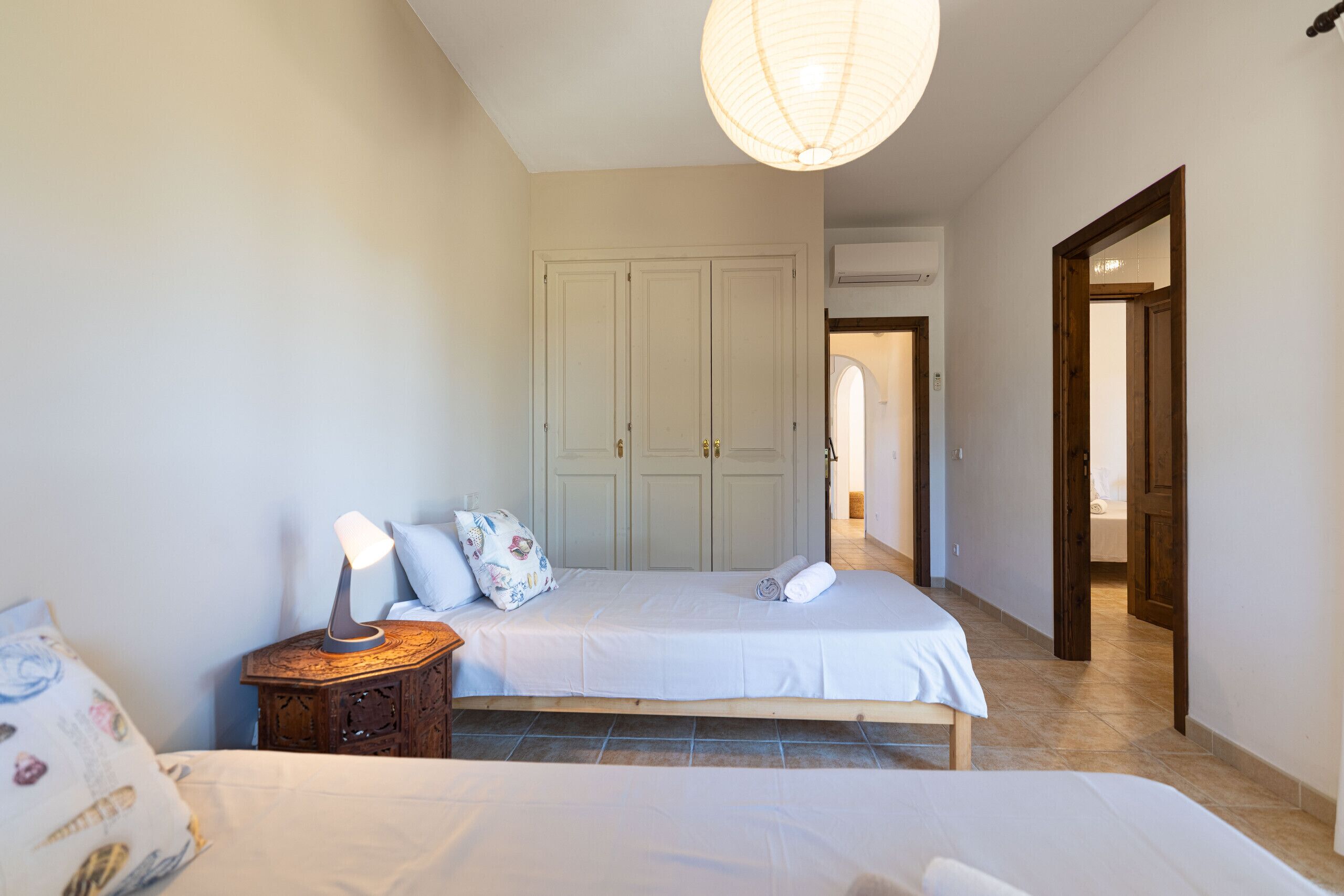 A bedroom with two beds, a bedside table with a lamp, a wardrobe, and a hallway leading to another room.
