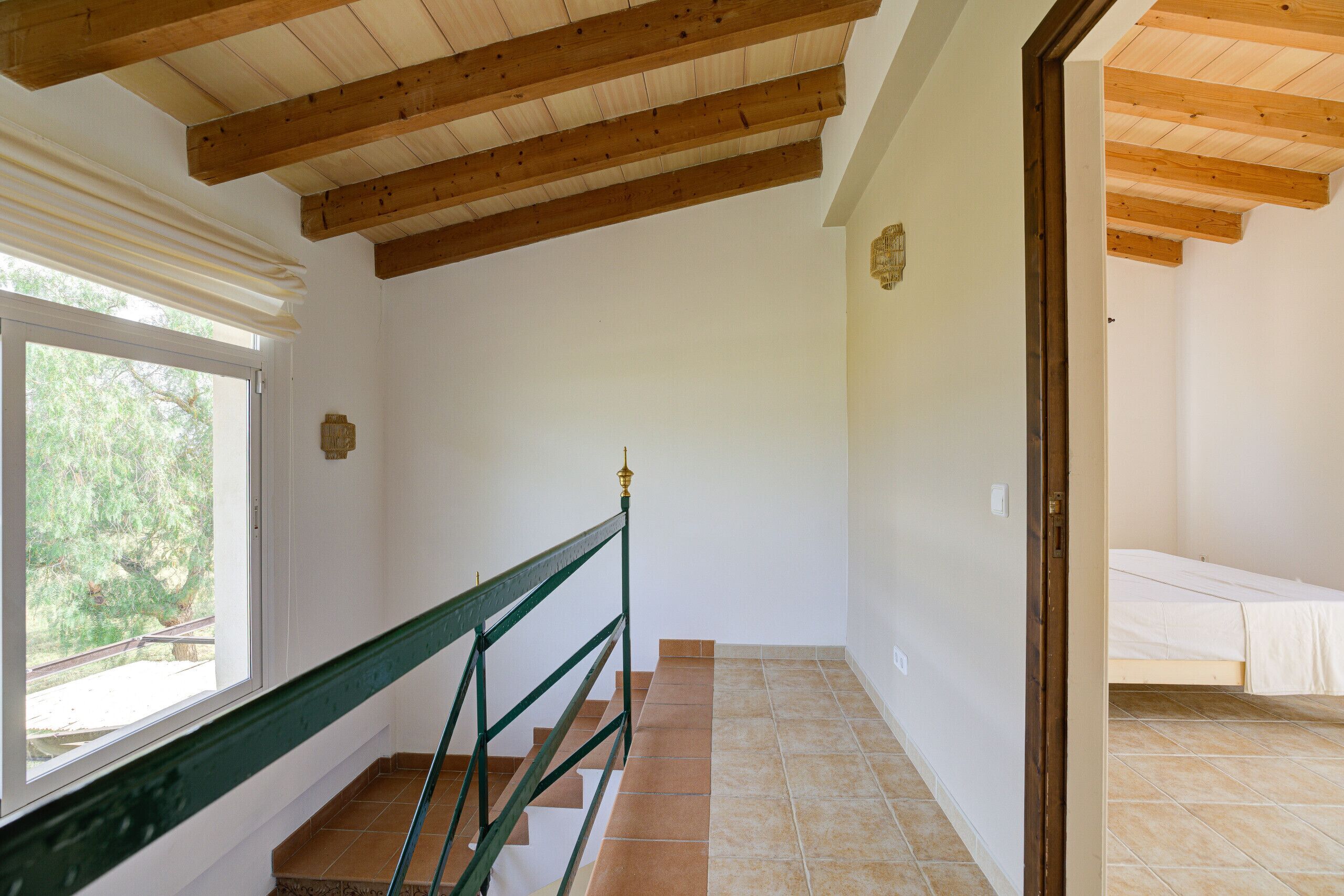 Una habitación con techo de madera, una ventana con persianas, una escalera con barandilla verde y una cama con colcha blanca.