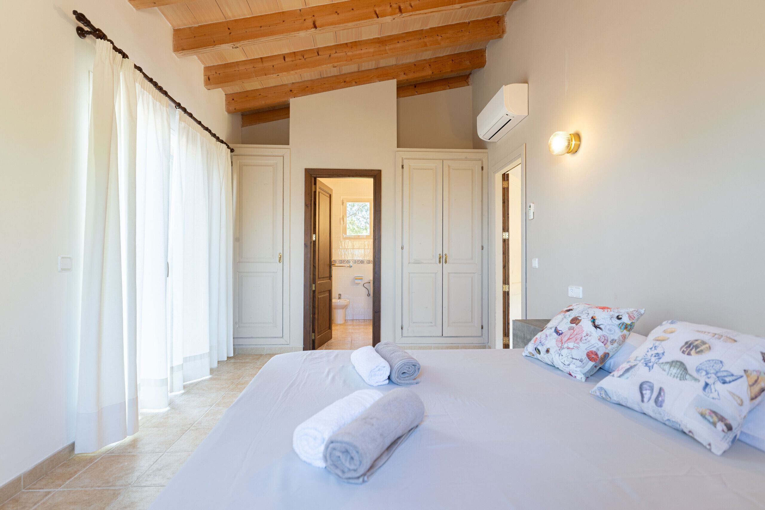 A bedroom with a bed, two pillows, a window with curtains, a door leading to a bathroom, and a ceiling with wooden beams.