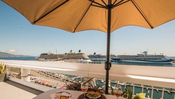 Uma mesa com comida e bebidas, um grande guarda-sol e vista para navios de cruzeiro no porto.