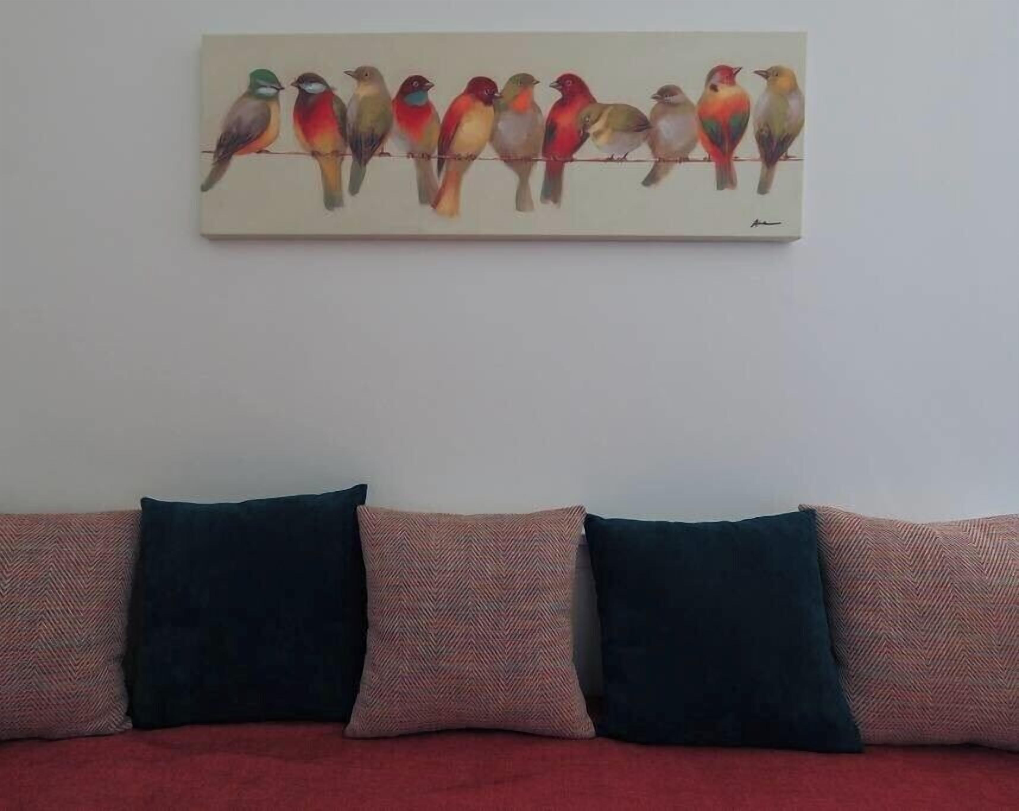 Une peinture représentant des oiseaux aux couleurs vives, accrochée au mur au-dessus d’un canapé agrémenté de coussins à motifs.