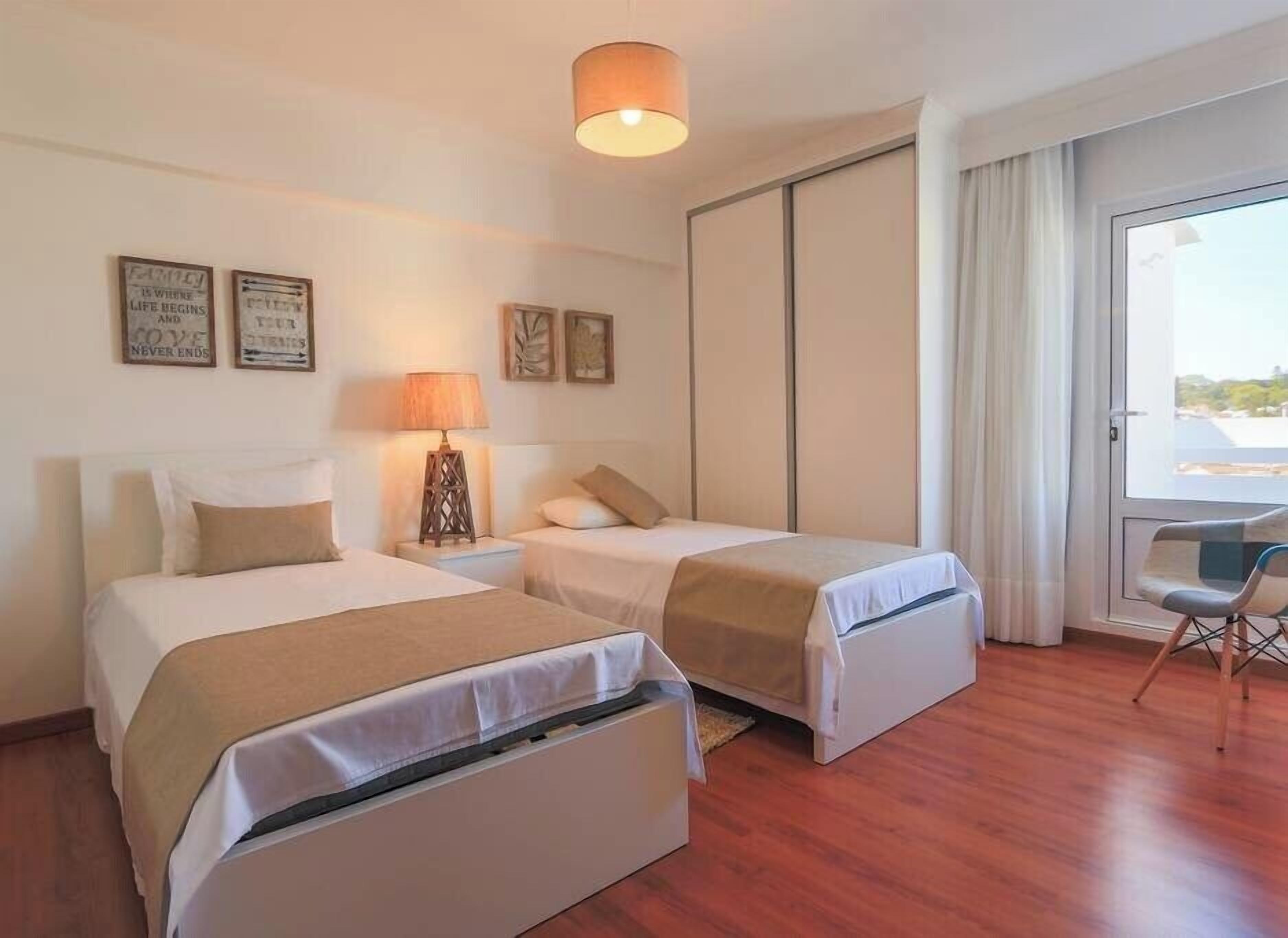 A hotel room with two beds, a desk, and a chair. There are two wall-mounted lamps and two framed pictures.