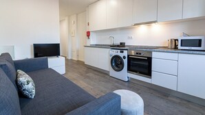 A modern kitchen with white cabinets, a washing machine, a microwave, and a grey sofa with a patterned pillow.