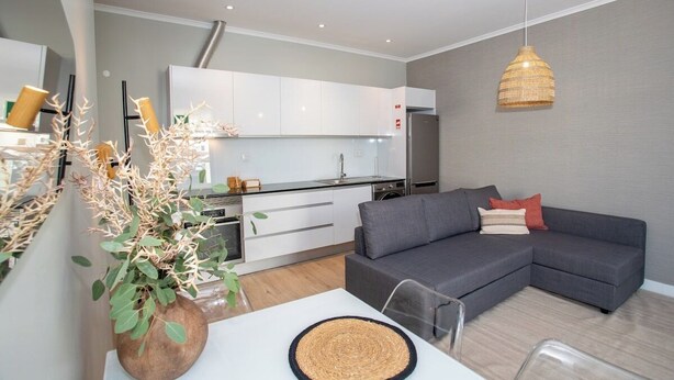 A modern living space with a grey sofa, a dining table with a woven placemat, and a kitchen area with white cabinets and a hanging light fixture.